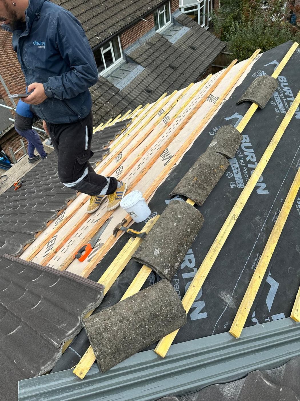 Roofer standing on new battens with hammer, snips and bucket, old mossy tiles set aside