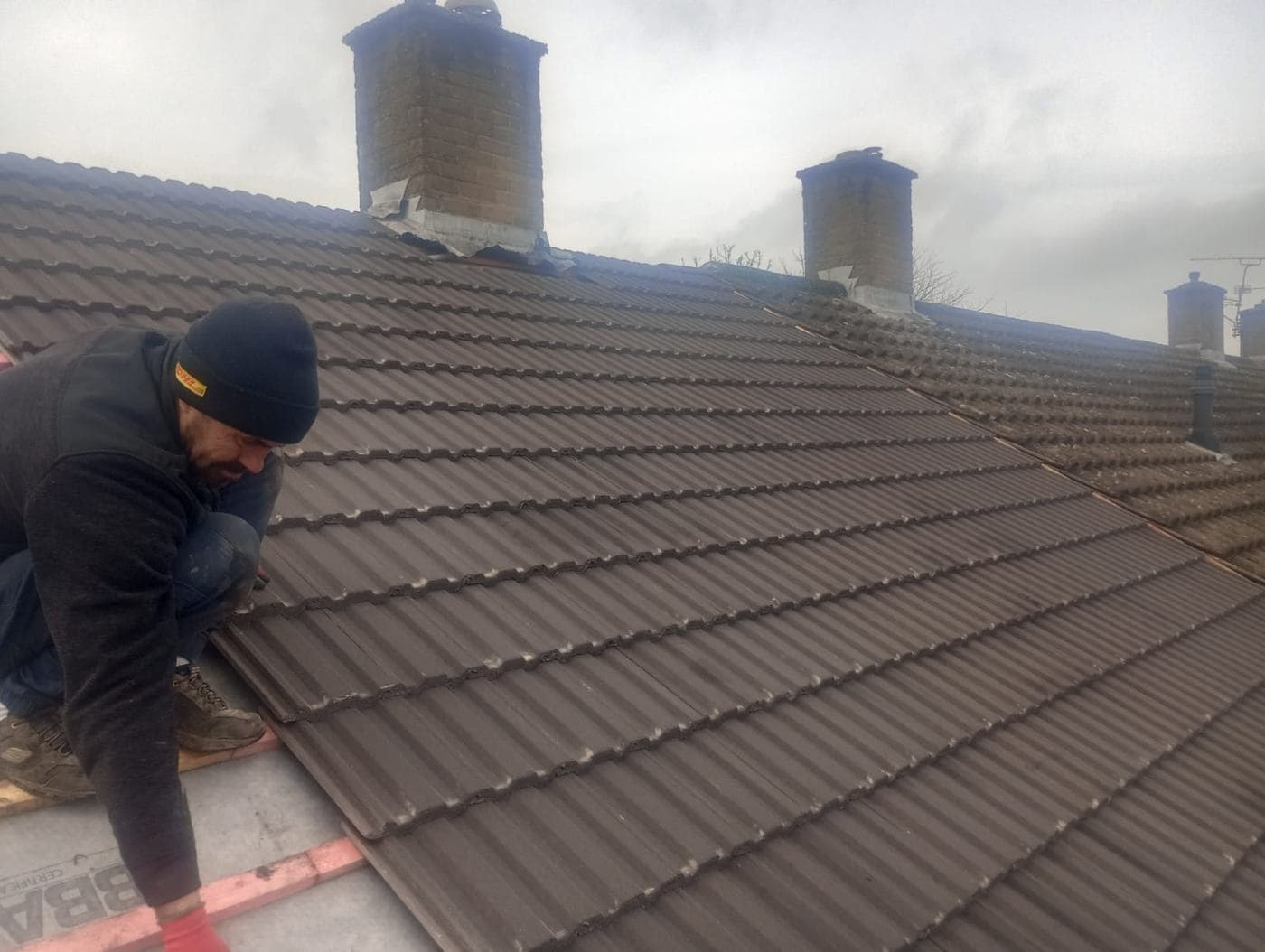 Roofer in a beanie working across dark brown tiles, brick chimney stacks behind