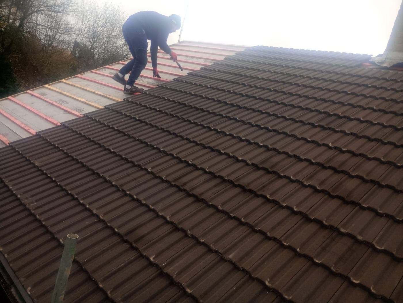 Roofer with a hammer tiling down the slope, marked battens and membrane still bare above
