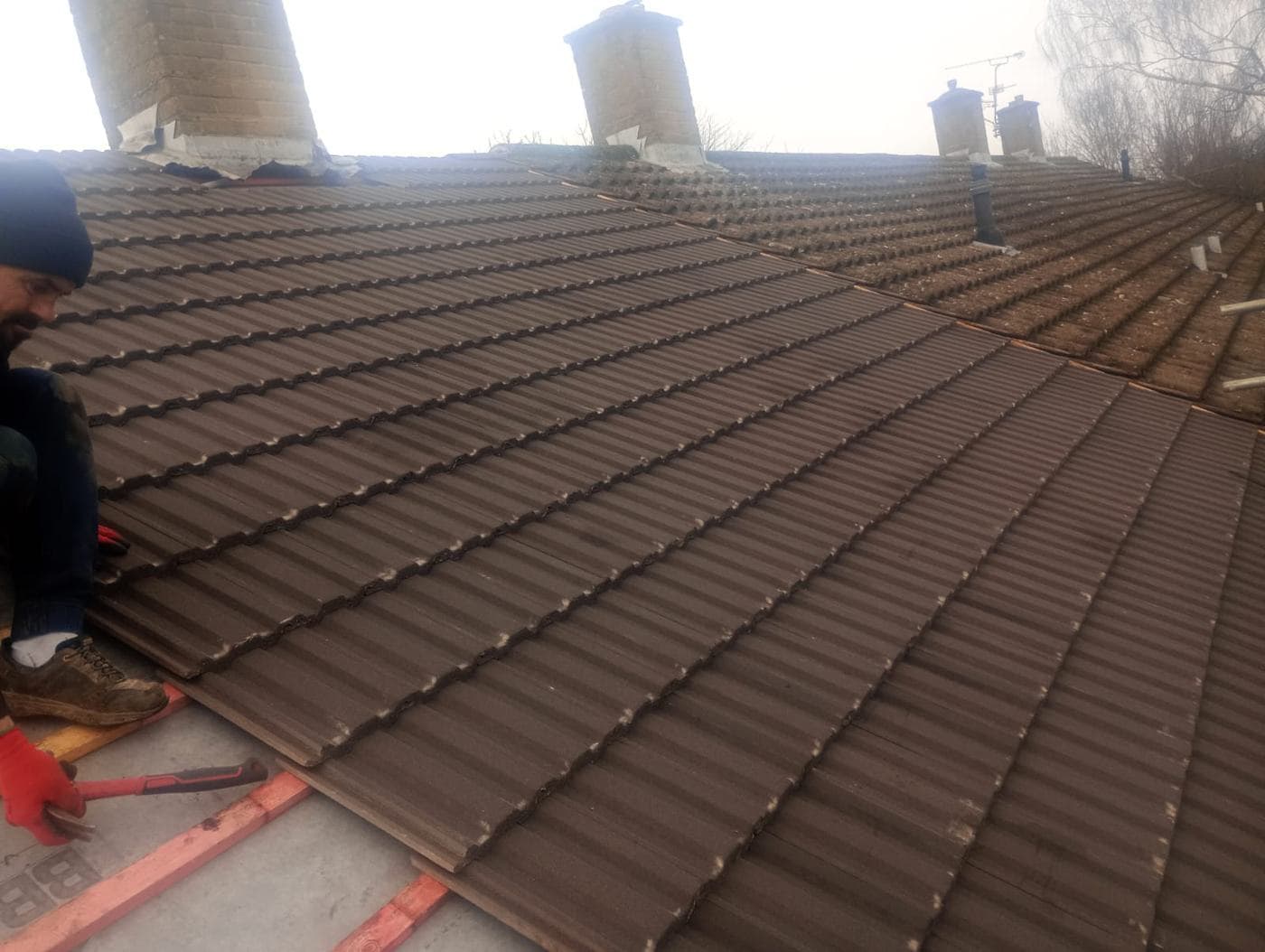 Roofer kneeling on the slope laying brown tiles, chimney stacks in winter mist