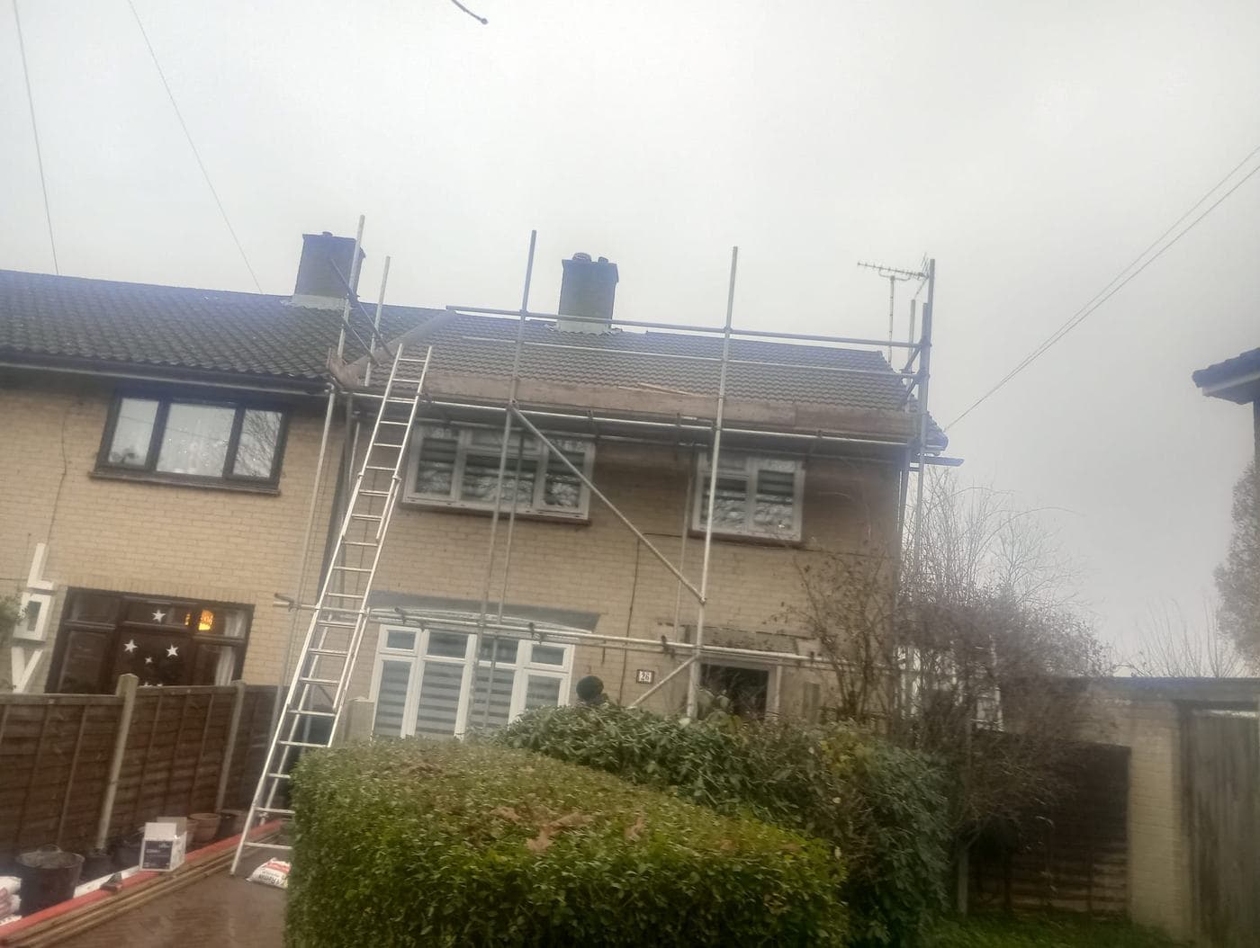 Scaffolding and a ladder against a cream brick house, roofline stripped back under grey sky