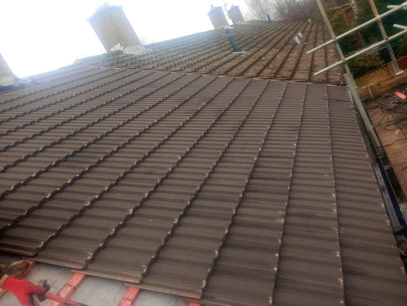 Long tiled slope rising towards pale chimney stacks, a roofer's boot in the foreground