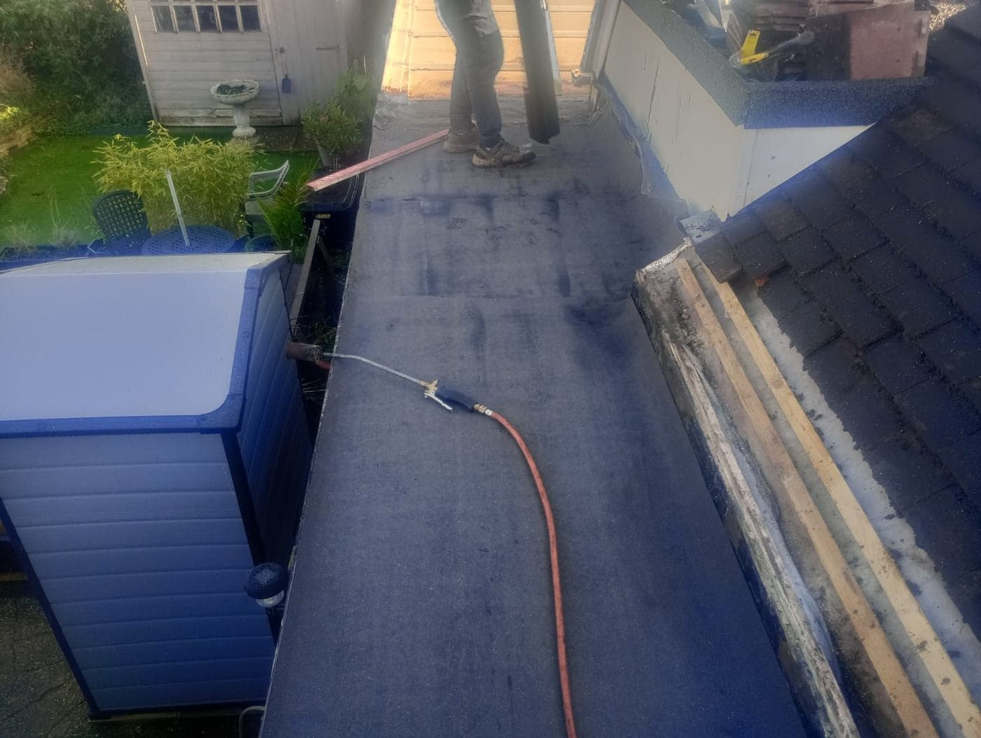Gas torch and hose resting on freshly bonded flat roof felt, garden below