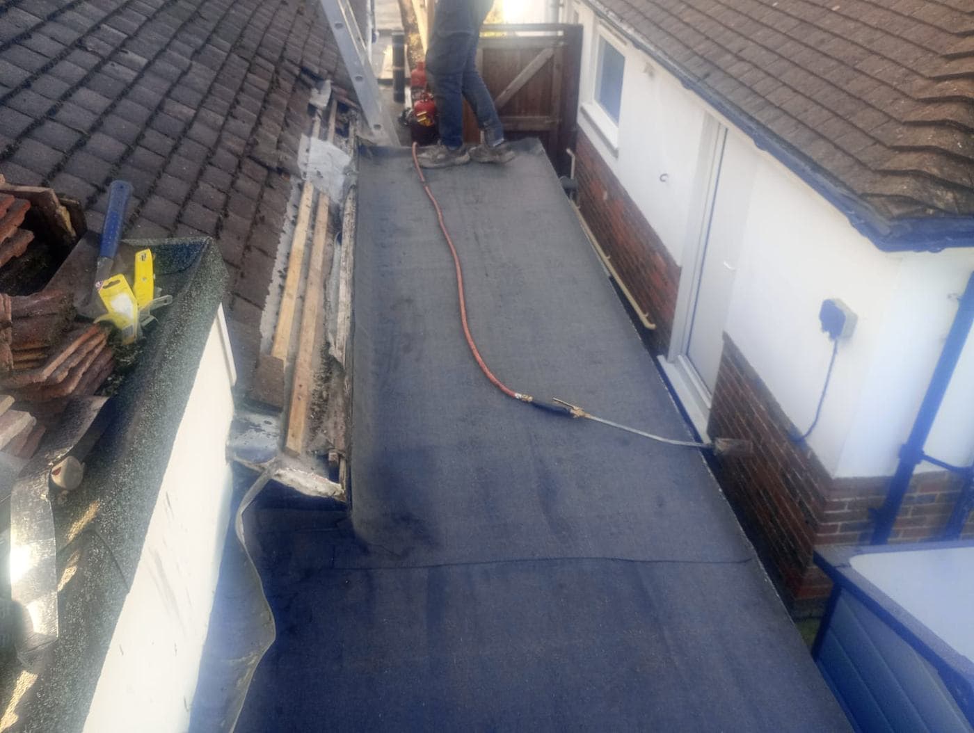 Roofer torching on flat roof felt, old clay tiles stacked beside stripped timbers