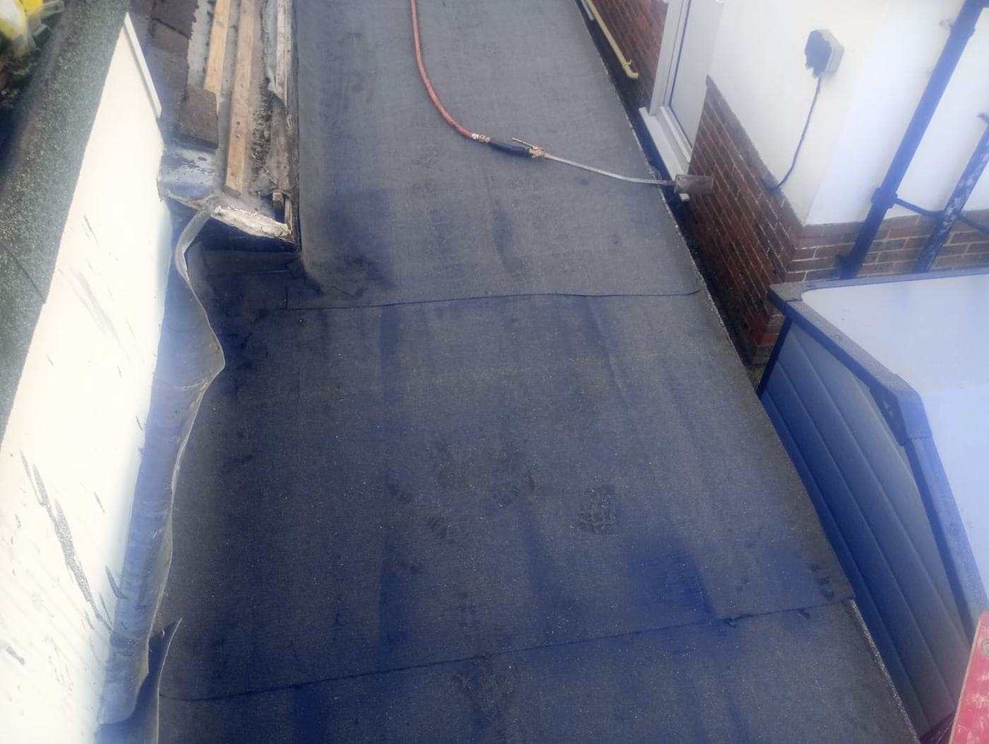 Torch-on felt flat roof between a rendered wall and red brickwork, gas torch resting on it
