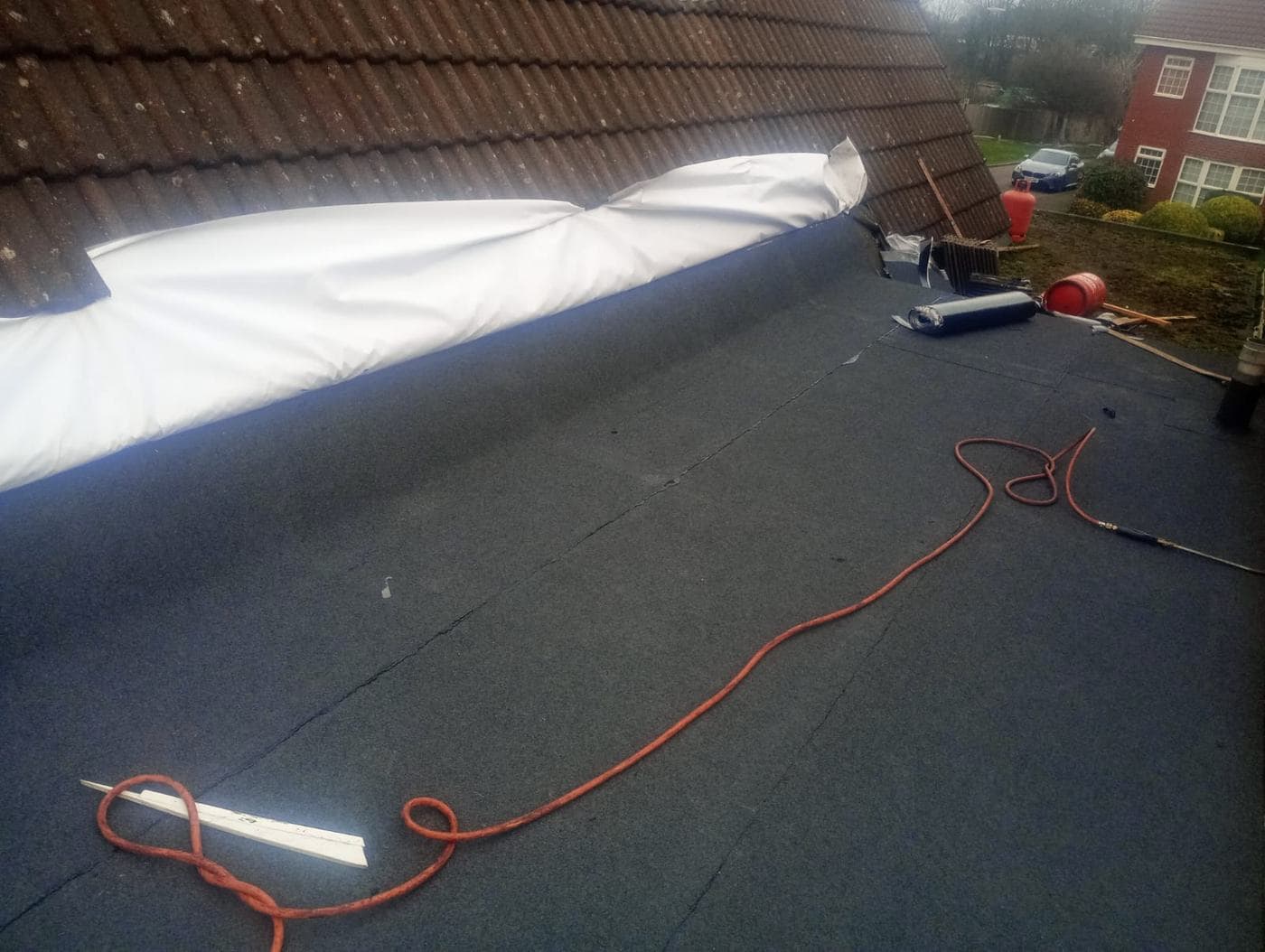 White protective sheeting draped over brown pitched tiles above freshly laid flat roof felt